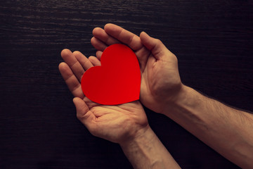 The outstretched arms on a background of brown wood texture. In the palms of your hands is a heart cut out of red paper.