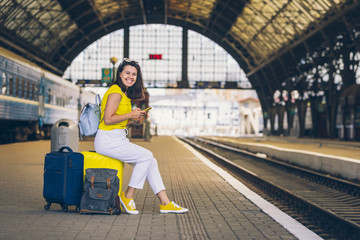 smiling pretty woman sitting on yellow suitcase with wheels at railway station