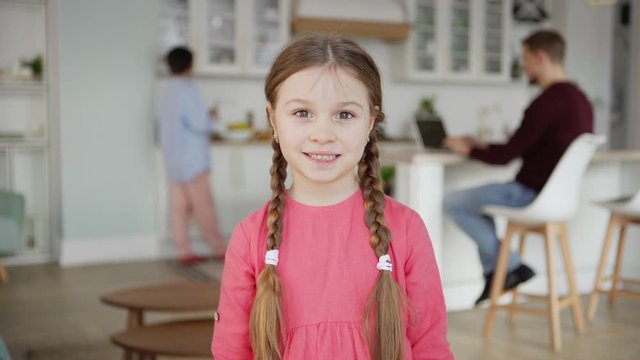 Waist up portrait of cute little girl with two braids looking at camera and smiling standing at home, her father working on laptop and mother washing fruits in background