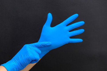 A nice girl or woman puts on a blue medical glove on her left hand. Only hands and gloves are visible. Close-up. Dark background. Copy space.