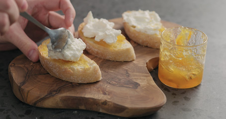 man spreading riccotta over orange marmalade on ciabatta slices on wood board