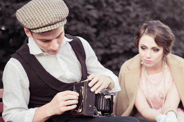 A young couple is photographed on a retro camera while walking in the Park. A woman sits on a bench and looks at the camera, a man opens the camera. historical reconstruction