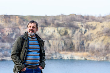 A middle-aged bearded man with a dark backpack stands on the shore of a flooded quarry.