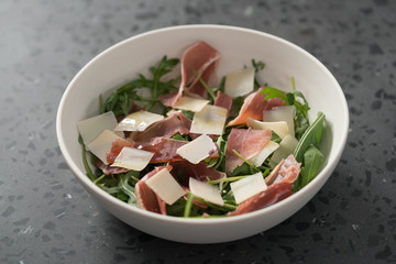Salad with prosciutto, parmesan and arugula in white bowl on concrete surface