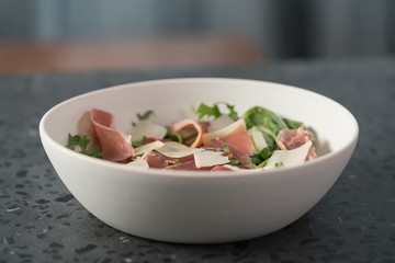 Salad with prosciutto, parmesan and arugula in white bowl on concrete surface