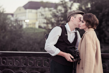 Fototapeta premium A young man and woman stand in a Park near a metal fence and hug, a man holding a retro camera in his hands.historical reconstruction