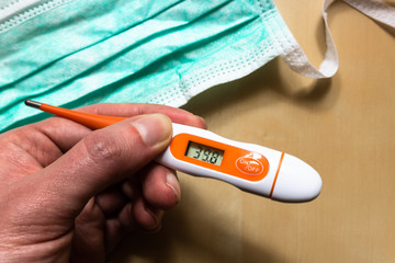 Hand holding digital thermometer displaying high temperature or fever reading with medical face mask in the background. Coronavirus COVID-19 pandemic background. Self-quarantine at home.