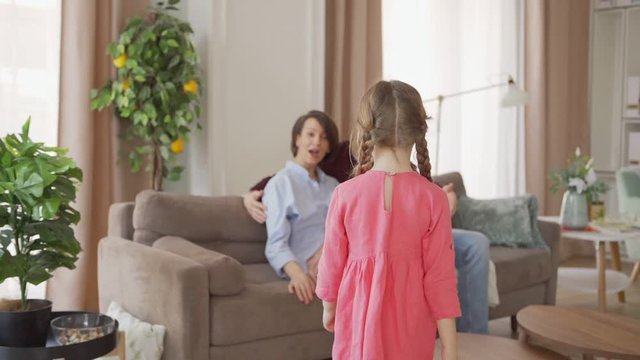 Follow slow motion shot of cute little girl walking to loving mother and father sitting on sofa at home. Happy parents enjoying talking cheerfully with adorable daughter sitting on mothers lap