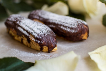 French eclairs with custard sprinkled with powdered sugar and white roses, petals, green leaves on light background