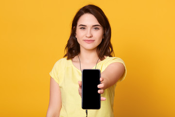 Indoor shot of young happy attractive european female with smartphone in hands, showing its bolank screen, posing isolated over yellow background, listening to music via earphones. Copy space.