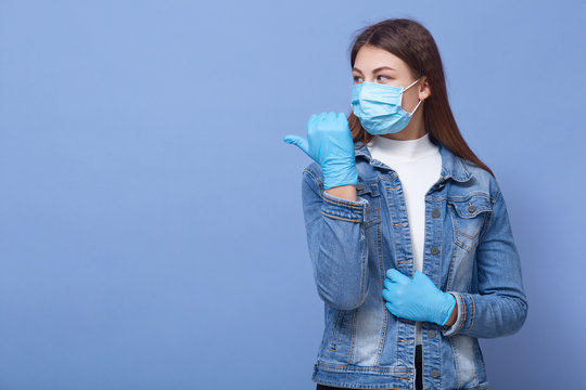 Close up portrait of beautiful and young woman wearing white shirt and denim jacket, blue rubber gloves and flumask on face, pointing aside with thumb, isolated over blue background, copy space.