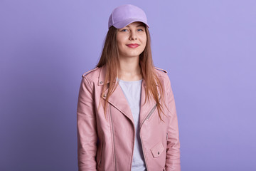 Horizontal indoor picture of cute lovely attractive teenager standing isolated over lilac background in studio, wearing cap, t shirt and leather jacket, looking directly at camera. Youth concept.