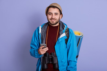 Close up portrait of young handsome men with backpack holding smart phone. Smiling student traveler portrait. Cheerful man posing isolated over lilac background. Lifestyle and people concept.