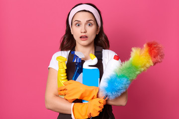 Photo of exhausted impressed housekeeper holding pp duster and detergents in different bottles, having shocked facial expression, opening mouth and eyes widely, hate job. People and clean up concept.
