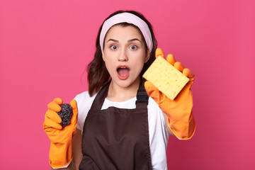 Close up portrait of young housewife being ready for cleaning, time for cleaning, astonished female with yellow sponge in hands looking at camera with opened mouth, wearing white shirt and apron.