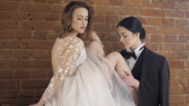 Wedding homosexual couple smiles together. Playful bride enjoy gentle touch of careeful female groom stroking her legs indoor. Intimate desire, hot romantic hug of two lovers feel seduction affection