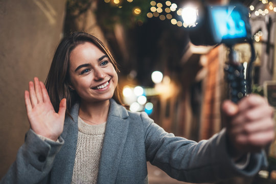 Traveler female blogger shooting video for social media with digital camera showing hi. Smiling woman vlogger taking photo selfie on background light night city illuminations