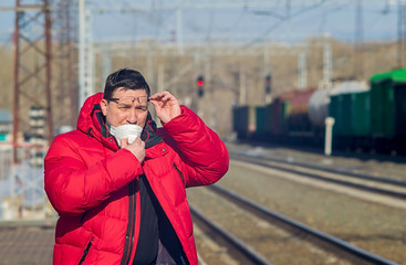 Fototapeta premium a man lifts his glasses and stands on the platform of a train station waiting for the train to arrive wearing a medical antiviral breathing mask