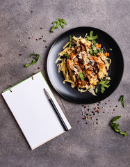 Bolognese pasta with mushrooms and arugula poured balsamic sauce on a black plate. A book for writing recipes lies on a table.
