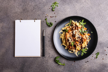 Bolognese pasta with mushrooms and arugula poured balsamic sauce on a black plate. A book for writing recipes lies on a table.
