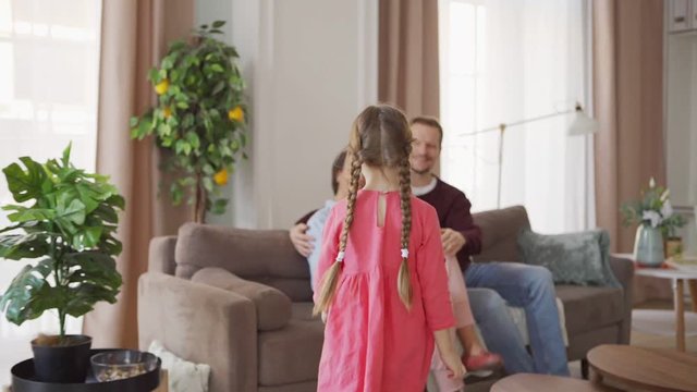 Follow slow motion shot of cute little girl with two braids walking to loving mother and father sitting on sofa at home. Middle aged parents enjoying talking with daughter sitting on mothers lap