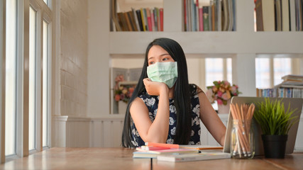 Young female students wear a mask and look out the window. She prepares her home exams to protect against the coronary virus or Covid-19.