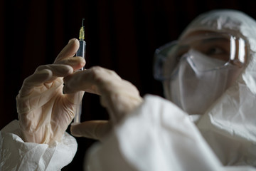 Woman wearing gloves with biohazard protective suit and mask holds an .virus vaccine with syringe,...