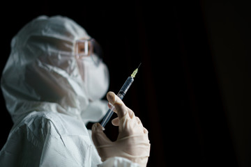 Woman wearing gloves with biohazard protective suit and mask holds an .virus vaccine with syringe,...