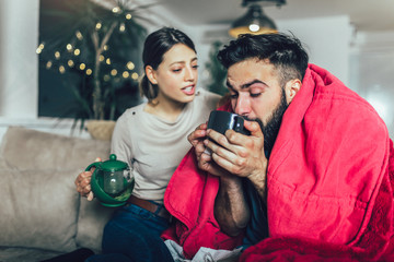 Cheerful romantic couple is sitting on sofa under cozy plaid and smiling. They are drinking tea.