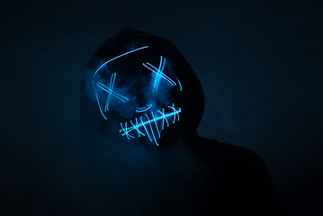 A man in a scary neon glowing mask and a hood on a dark background with smoke. Horror and Halloween...