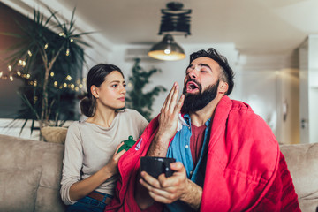 Cheerful romantic couple is sitting on sofa under cozy plaid and smiling. They are drinking tea.