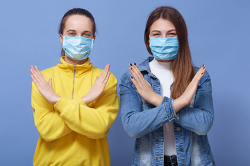 Indoor shot of two young woman wearing bright clothing and medical disposable masks, showing stop gesture with hands isoalted over blue background. Flu, coronavirus or contagious diseases concept.