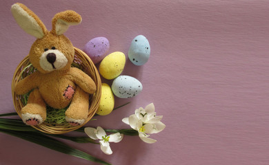 Easter holiday flat lay. Fluffy toy rabbit in a nest with flowers and multicolored eggs on a lilac, purple, violet background. Copy space for your text