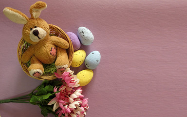 Easter holiday flat lay. Fluffy toy rabbit in a nest with flowers and multicolored eggs on a lilac, purple, violet background. Copy space for your text
