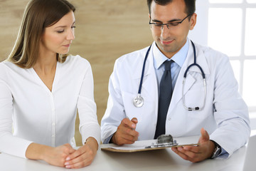 Doctor and patient discussing the results of a physical examination while sitting at a desk in a clinic. A male doctor using a clipboard to fill out a medical history of a young woman's medication