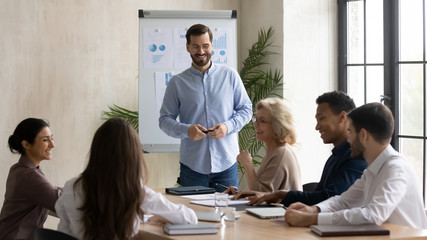 Smiling male coach joke make whiteboard presentation for multiracial businesspeople at briefing, happy diverse colleagues have fun laugh discussing business ideas brainstorming at team meeting