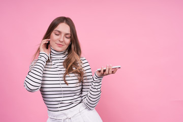 Fototapeta premium Young emotional caucasian girl in headphones and a phone on a pink background.