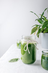 Three glass jars with spinach shake on a table with a white tablecloth and a plant