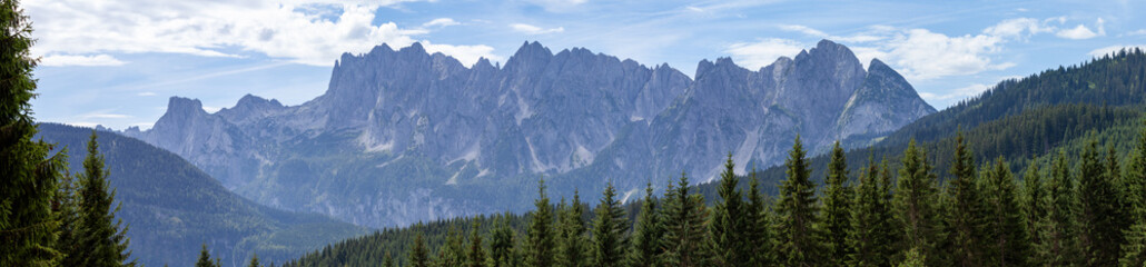 Famous Gosaukamm in Upper Austria. Seen here in beautiful sunny summer weather with some clouds in the blue sky. Green pine trees are all over the mountains in front.