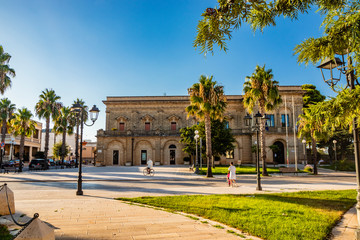 Fototapeta na wymiar Acquarica del Capo, Presicce, Salento, Puglia, Italy. Town square with the seat of the municipality and the town hall. The ancient stone and brick building. Flower beds with the lawn and palm trees.