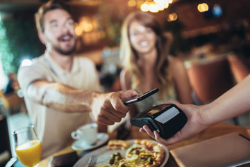 Happy couple using mobile phone and making contactless payment in a restaurant. S