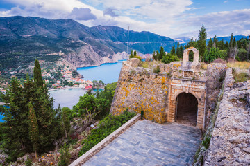 Fototapeta na wymiar Old fort gate on top of frourio peninsular and Assos village with beautiful sea bay and cypress trees in background. Kefalonia island, Greece