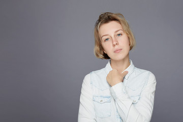 Charming young pure and innocent Caucasian woman has fresh skin, looks at camera with dreamful face expression, keeps hand on denim blue shirt,  being deep in thoughts, posing indoors on grey wall .