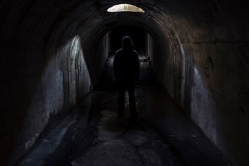 A figure of a man in a hood walking forward in a dark underground tunnel lit through a hatch in the ceiling. Fort Pospelova, Vladivostok.