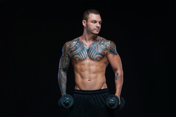 Fototapeta na wymiar Tattooed strong muscular athletic man pumping up muscles with dumbbells on black background
