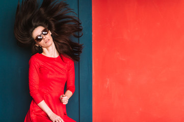 Beautiful fashion girl with long hair, spanish appearance in sunglasses and red elegnat dress posing on blue red wall in studio. Stylish brunette babe with flying hair. Professional hairstyle concept.