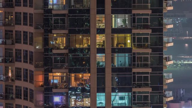 Rows of glowing windows with people in the interior of apartment building at night. Modern skyscraper with glass surface. Concept for business and modern life. Zoom out