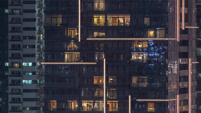 Rows of glowing windows with people in the interior of apartment buildings at night. Modern skyscrapers with glass surface. Concept for business and modern life. Pan left