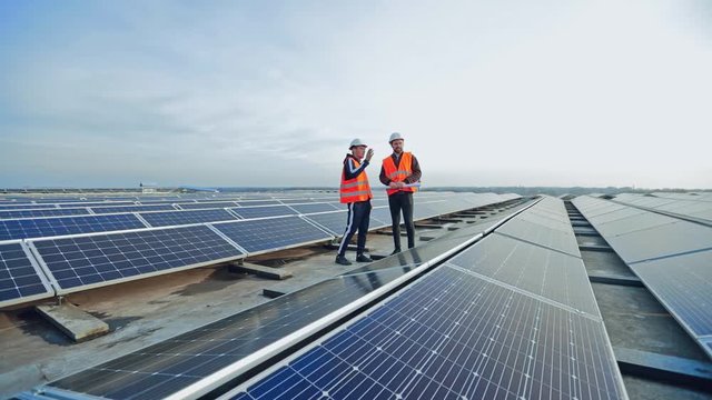 Modern solar farm on a roof under the sky. Workers in uniform and protective helmets discuss the solar panels installation.