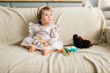 Obraz na płótnie Canvas Little cute girl in beautiful dress sits on the couch and plays with her various toys